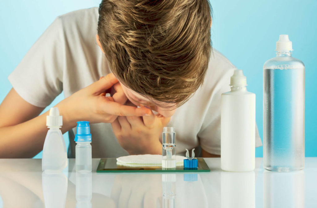 A boy puts on night contact lenses at the table with bottles of cleansing solution and eye drops.