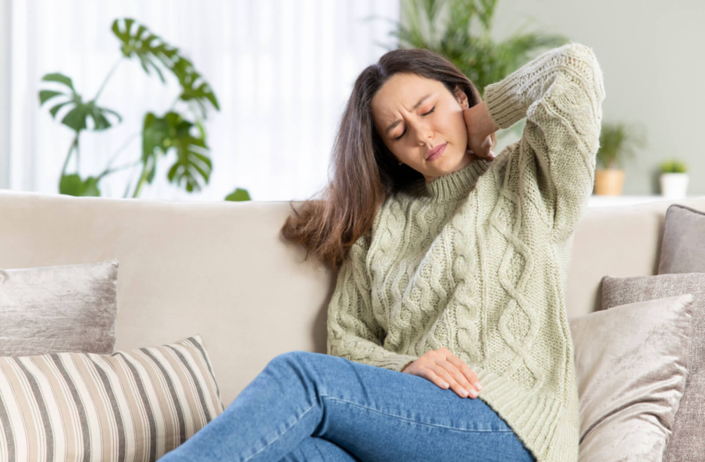 A woman a knitted green sweater experiencing neck pain while trying to relax in her living room.