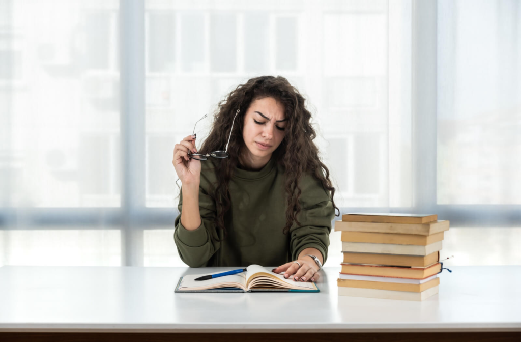 A young woman with a stack of books beside her showing signs of frustration while reading.