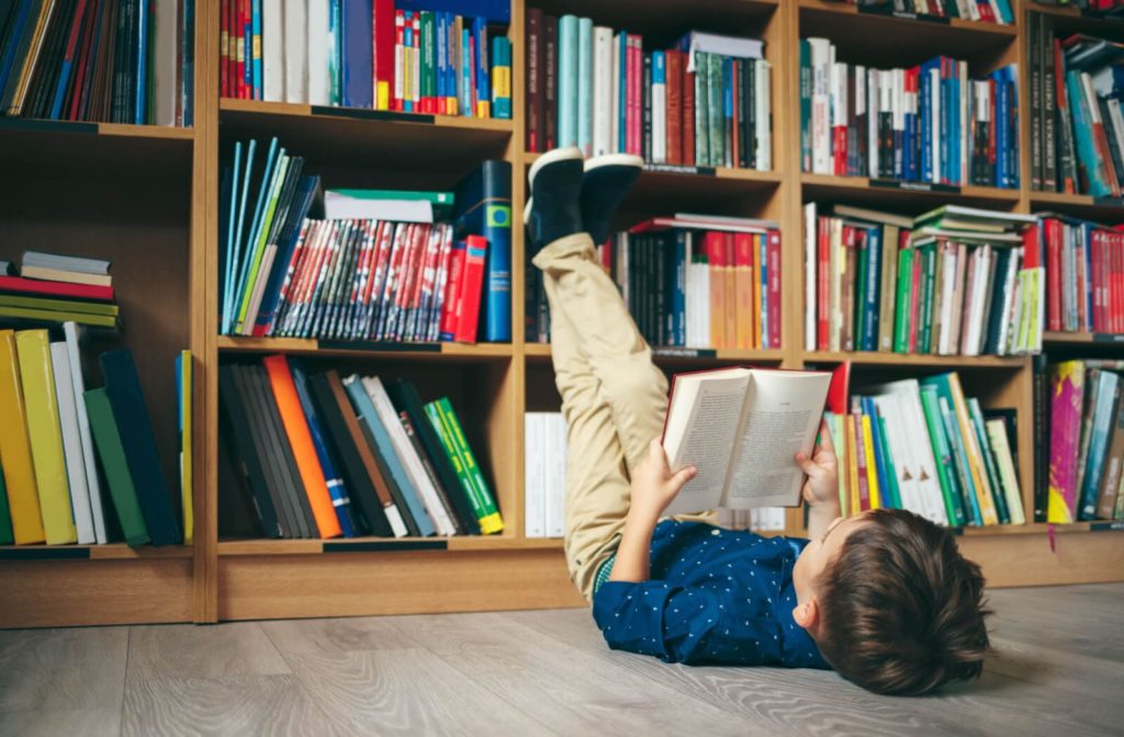 A young boy comfortably reading while lying down on the floor and resting his feet against a bookshelf full of books.