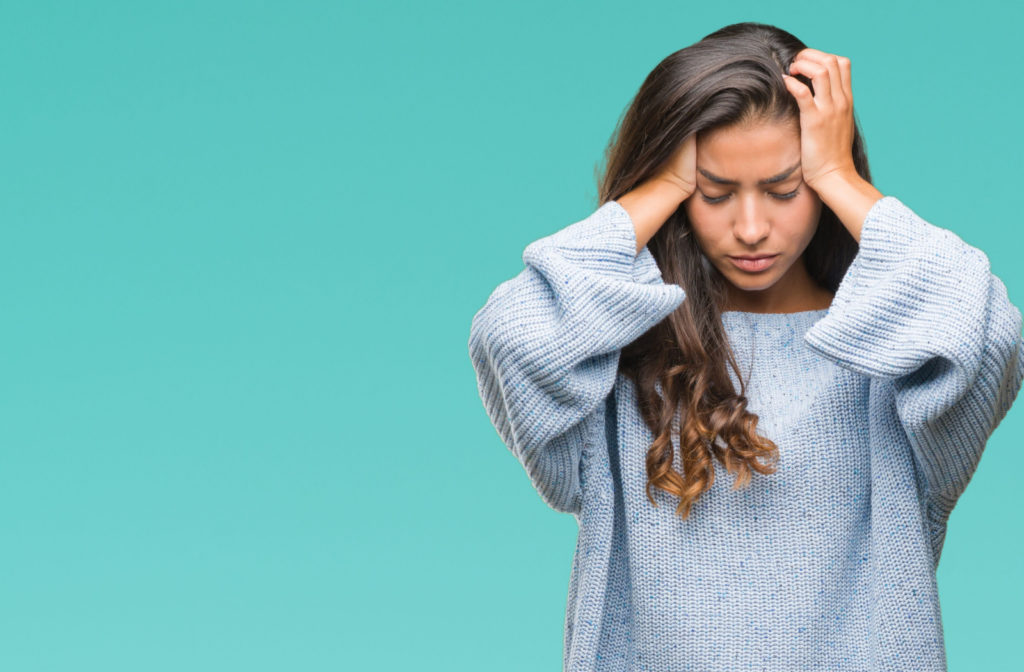 A stressed woman against a teal background with both of her hands applying pressure to her head.