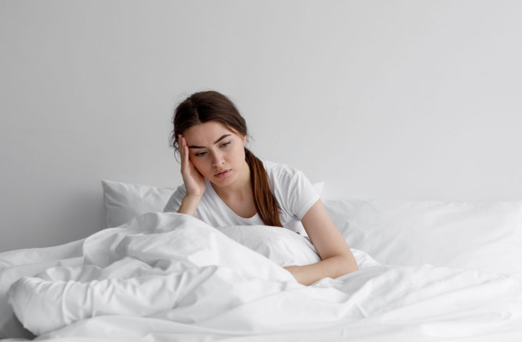 A woman sits up in bed, looking tired and frustrated with her head resting in her hand, as she is unable to sleep.