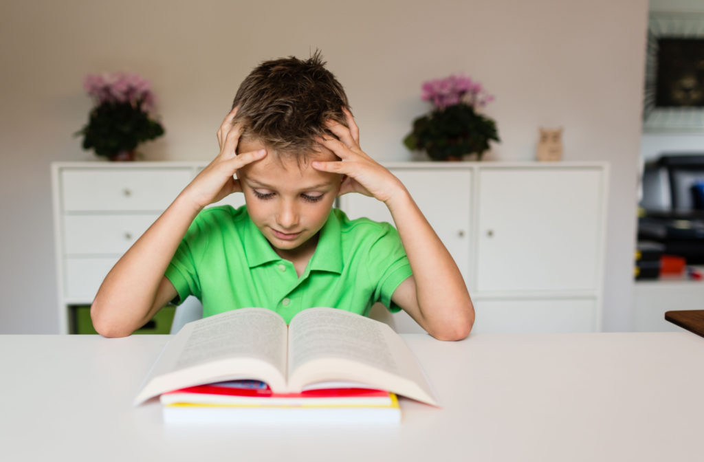 A young boy in a green shirt sitting at a desk and holding his hands to his head as he looks at an open book.