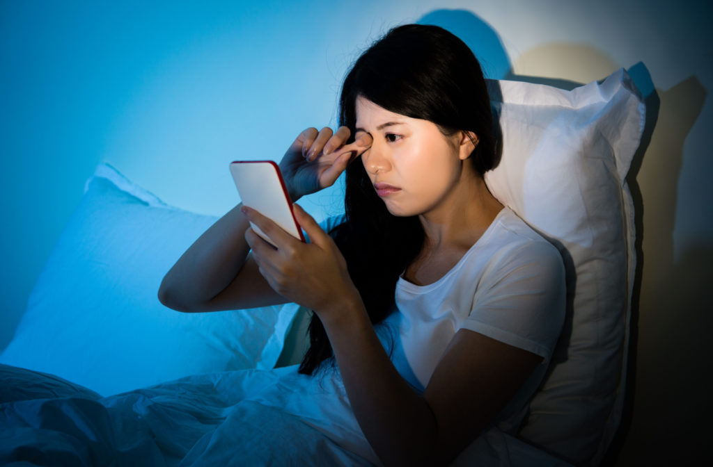 A woman rubbing her eyes while using her phone in a dark room.